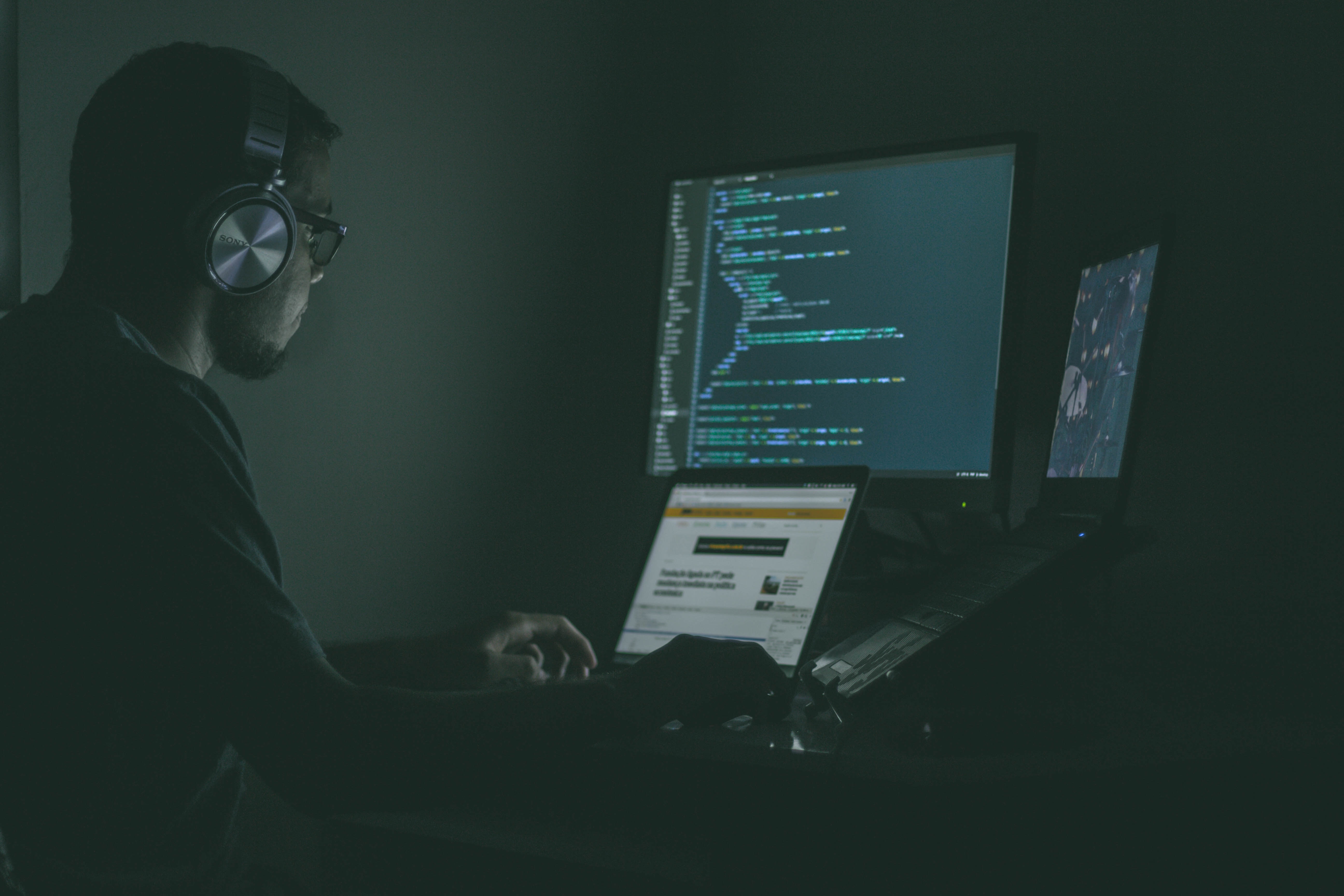 A man sits in a shadowy room in front of multiple computer screens. One is running a coding language.